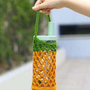Hand holding a glass water bottle wrapped in a green and orange crochet netting.