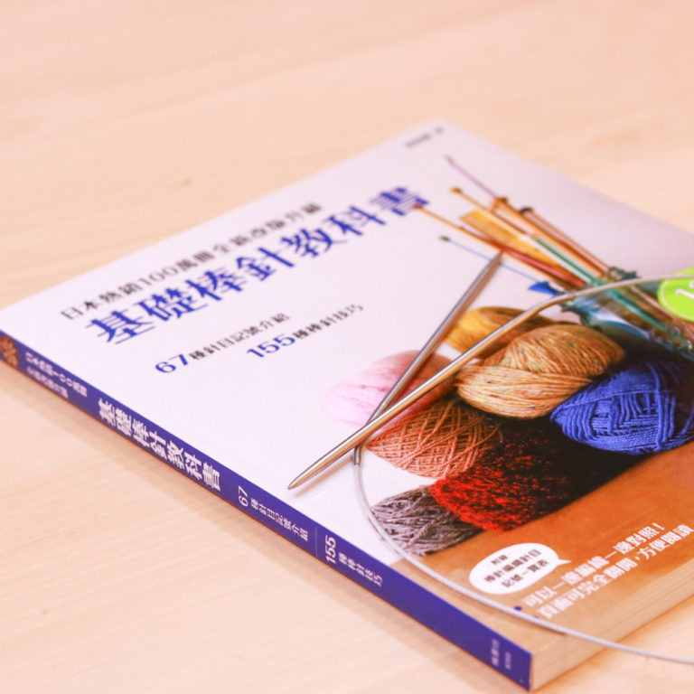 一本名為《基礎棒針教科書》的日系編織教學書，平放在木紋桌面上，書的封面上疊放著一副金屬棒針。