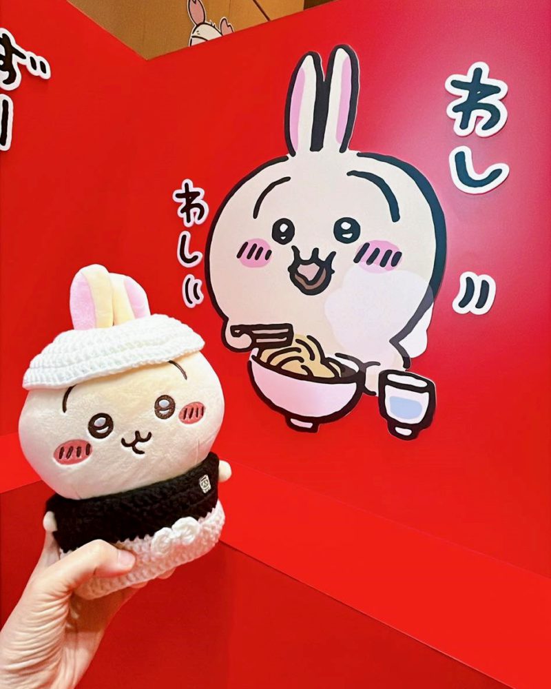 一隻Usagi絨毛玩偶，穿著一套鉤織的拉麵師傅服裝（白色帽子和黑色上衣），被舉在一面有著吉伊卡哇卡通圖案的紅色牆壁前。