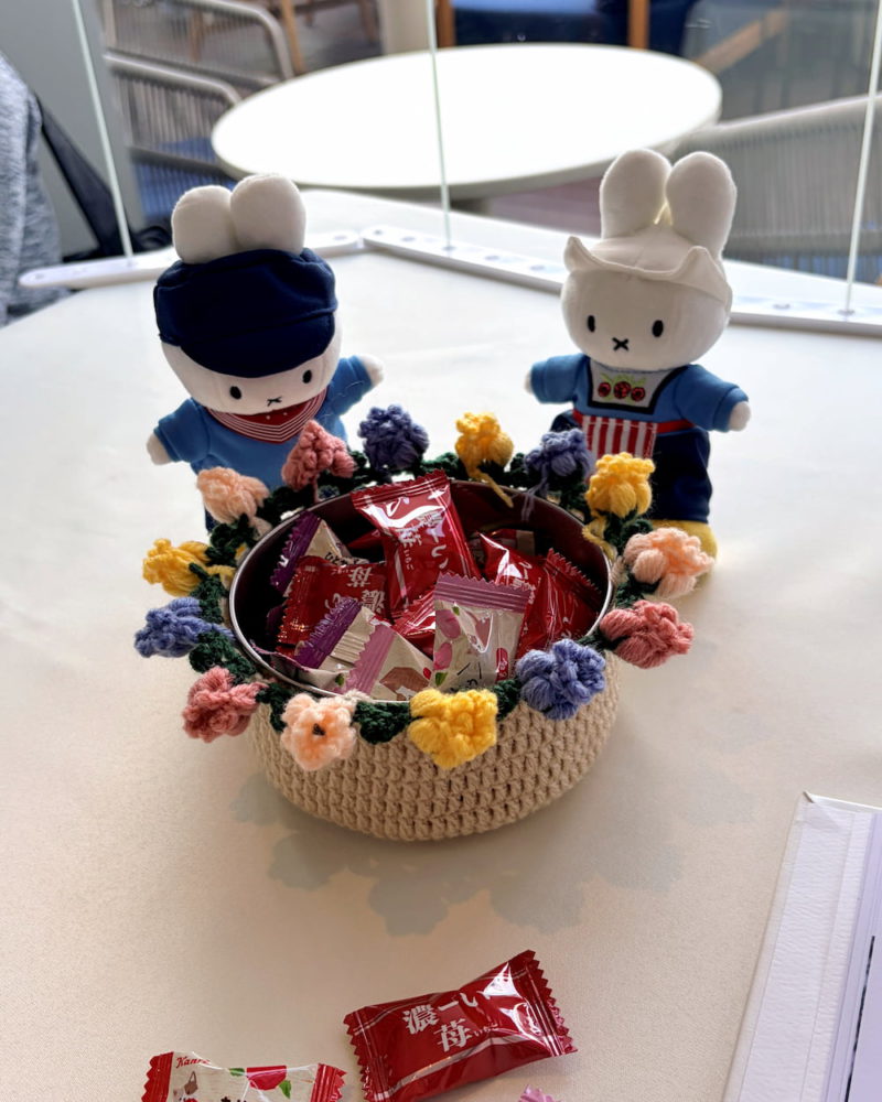 一個裝滿喜糖的圓形鐵罐，外面套著由Heyman鉤織的米白色針織套，罐口裝飾著一圈立體的彩色鈴蘭花朵，後方有兩隻穿著圍裙的Miffy玩偶。