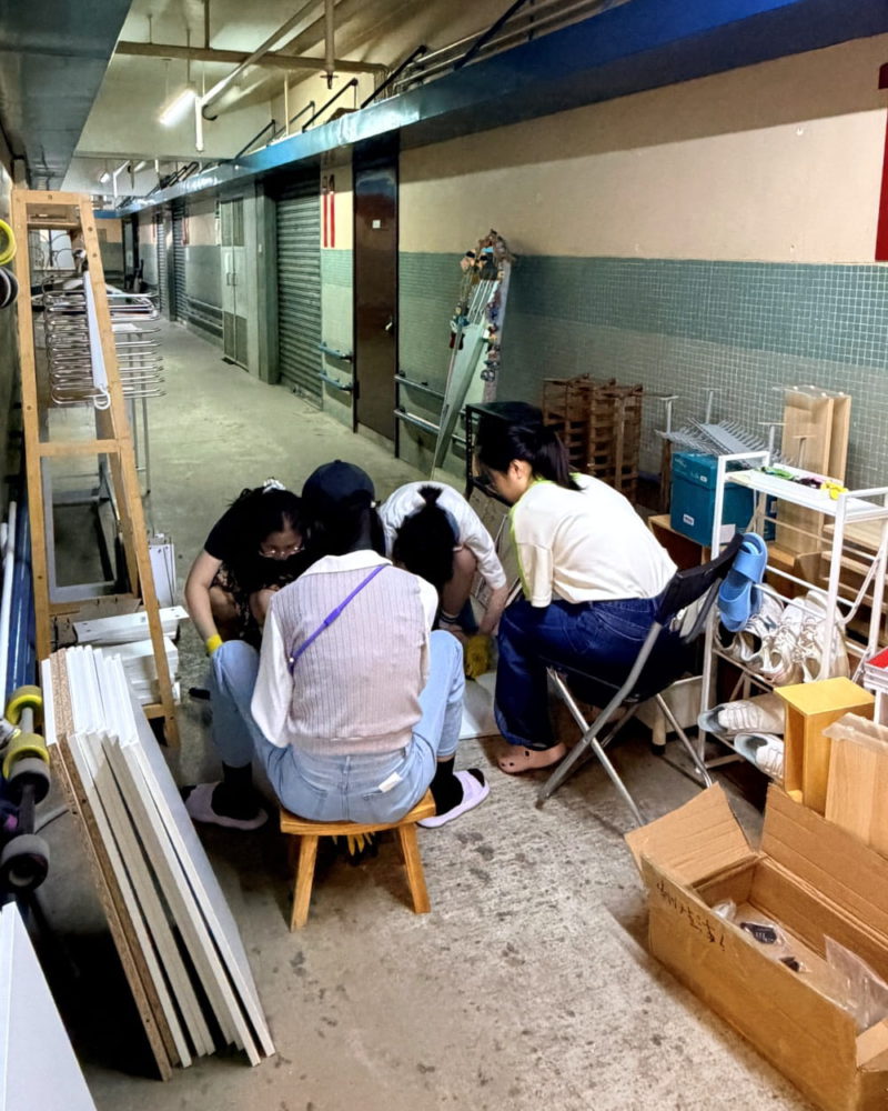 幾位女生蹲在昏暗的走廊地上，專注地組裝白色的木製傢俱配件，旁邊堆放著紙箱和工具，展現工作室裝修期間的忙碌。