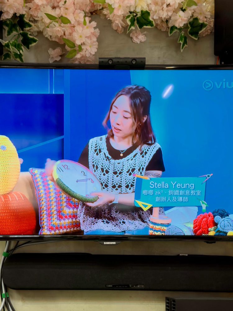 電視螢幕播放著ViuTV《智富通》節目，畫面中唧唧主理人Stella正在展示一個色彩繽紛的鉤織時鐘作品，桌上擺滿了各式鉤織公仔。