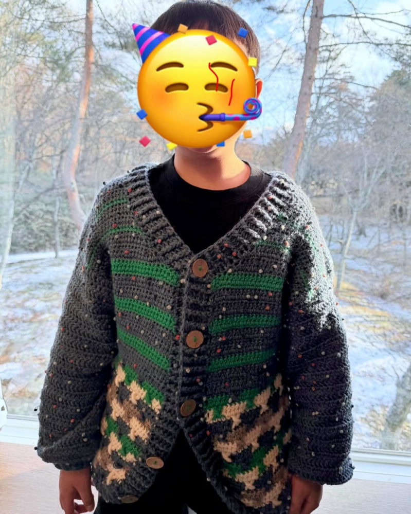 Remmy的兒子穿著一件以深藍灰色為主、帶有綠色條紋和底部幾何圖案的鉤織開襟外套，外套配有木製鈕扣。