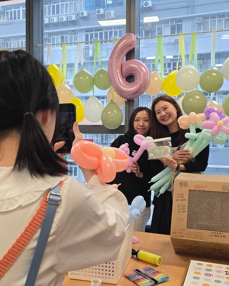 兩位女同學拿著色彩繽紛的造型氣球和唧唧六週年紀念品，在掛滿綠色與黃色氣球、並帶有數字「6」鋁箔氣球的落地窗前開心合影，前方由助教拿著手機為她們拍照。