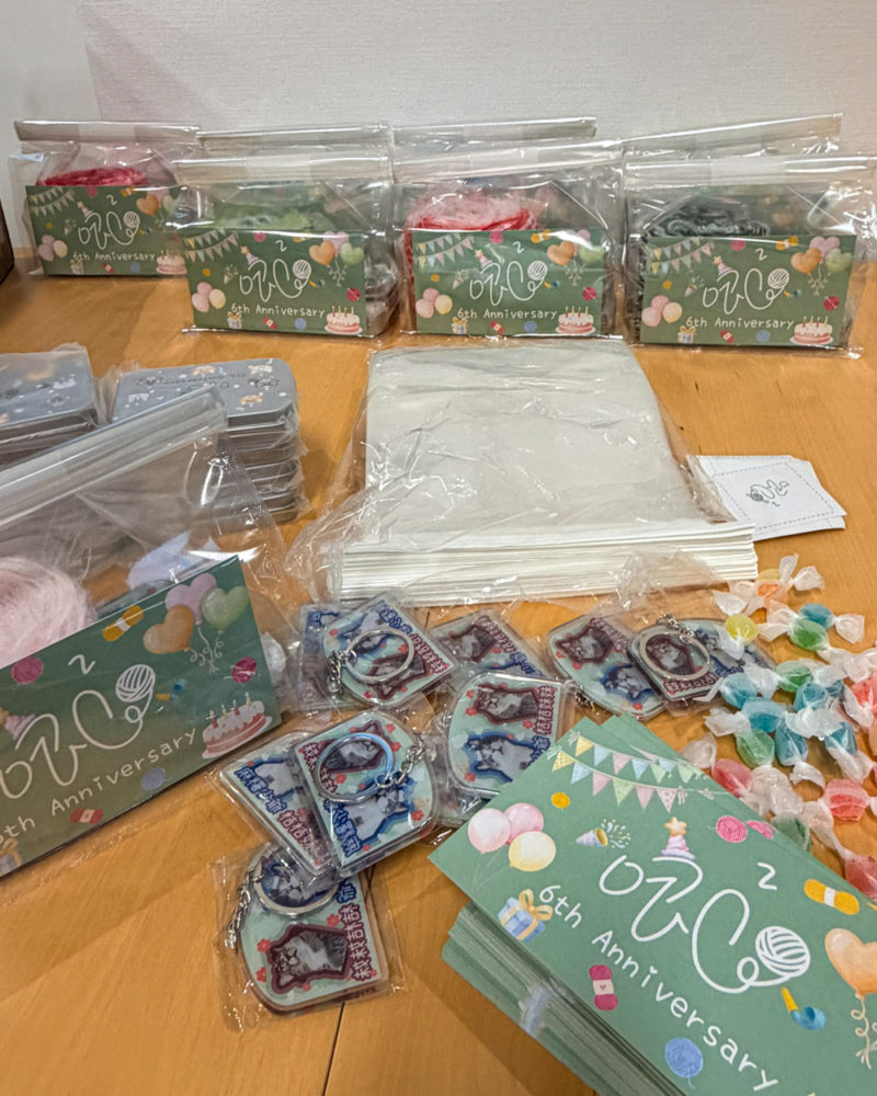 木桌上擺滿了唧唧準備的六週年紀念禮物，包含印有「zik2 6th Anniversary」的綠色包裝袋、手工繞製的手混線、裝在透明袋裡的店貓造型壓克力鑰匙圈，以及散落的彩色糖果。