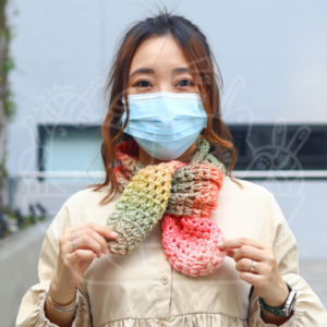 Woman wearing a blue surgical mask and a multicolored knitted scarf outdoors, facing the camera.