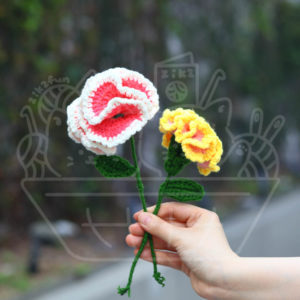 Two crocheted yarn flowers (pink and white, and yellow) with green stems being held by a hand in an outdoor street setting.