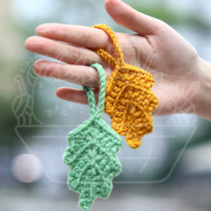 Hand holding two crocheted leaf ornaments, one green and one yellow, between the fingers.