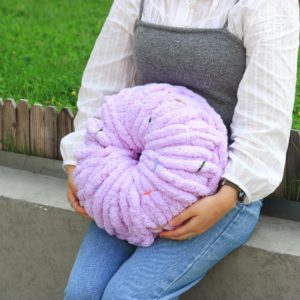 Person in a white shirt and gray top holds a large lavender chunky yarn ball outdoors by a bench/green grass.