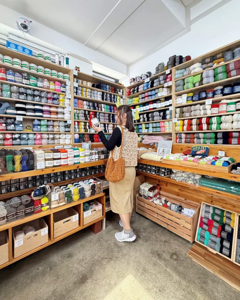 Woman browsing colorful yarn shelves in a yarn shop, holding a white skein and wearing a tan skirt and woven bag.