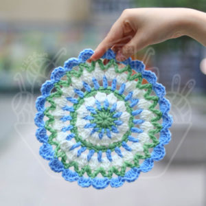Hand holds a blue‑green‑white crocheted mandala doily against a blurred outdoor background.