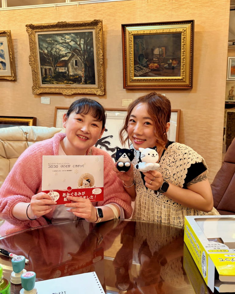 HAMANAKA社長與唧唧老師Stella坐在私人藝術品收藏室內合照，Stella拿着Mana、Yu主題繪本及黑白兩隻吉祥物玩偶，背後牆上掛有多幅畫作。