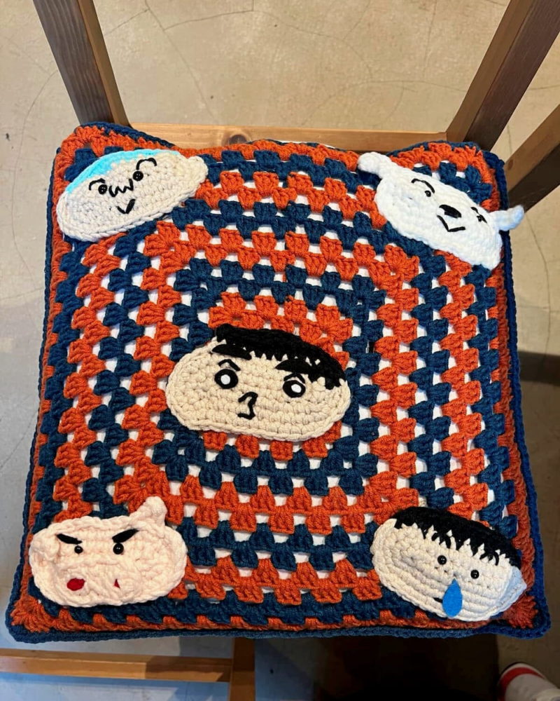Crocheted square pillow with orange and navy squares and four cartoon faces at the edges, each with simple expressions.