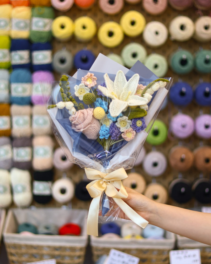 Hand holding a bouquet of knitted crochet flowers wrapped in plastic with a cream ribbon, in a yarn shop background with colorful skeins.