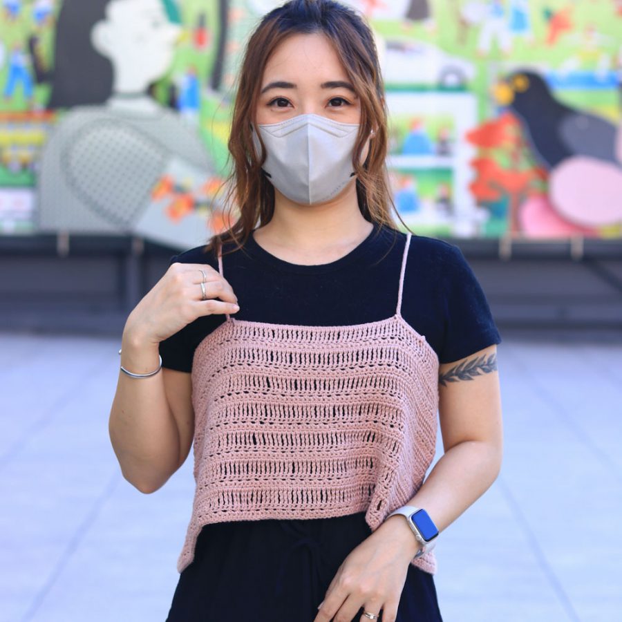 Woman wearing a gray face mask and pink crochet top standing outdoors in front of a colorful mural.